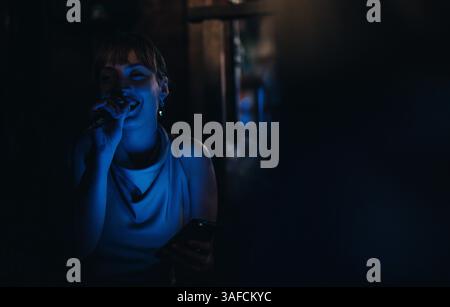Junge Frau, die leidenschaftlich in ein Mikrofon in schwach beleuchteter Atmosphäre singt, vermittelt Emotionen und Engagement in einer gemütlichen Umgebung, ideal für das Nachtleben Stockfoto