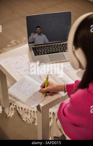 Mädchen, das Kopfhörer trägt und Notizen im Notebook macht, während sie während des Online-Kurses männlichen Lehrer auf einem Laptop beobachtet. Fernunterricht in Englisch Stockfoto