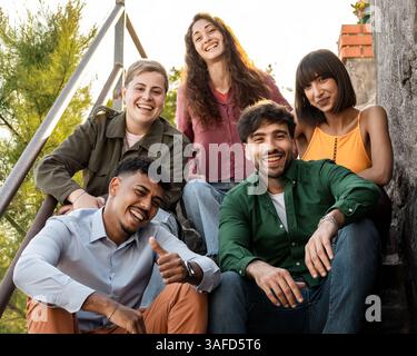 Eine vielfältige Gruppe von Freunden lächelt und posiert vor der Kamera auf den Stufen im Freien und genießt einen fröhlichen Moment bei natürlichem Sonnenlicht. Stockfoto