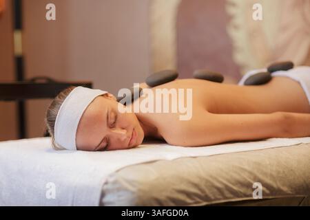 Junge Frau, die eine Massage mit heißem Stein in einem luxuriösen Spa erhält und entspannt auf einem Massagetisch im Raum mit Kerzenlicht liegt. Stockfoto
