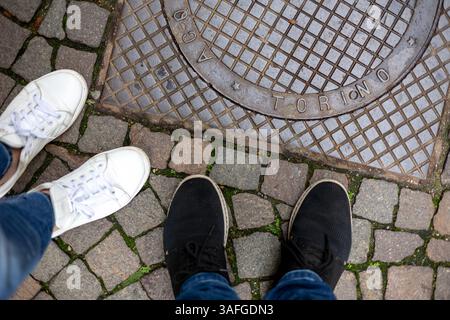 Ansicht von oben nach unten von zwei Paar Füßen in Turnschuhen, die auf Kopfsteinpflaster neben einer Metallabdeckung mit der Aufschrift „TORINO“ stehen. Stockfoto