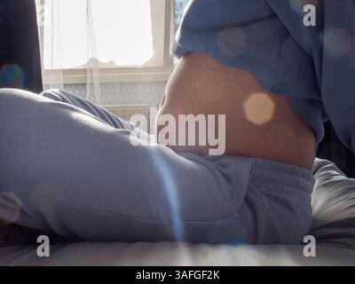 Schwangere Frau, die sich auf dem Bett entspannt, sanft den abgerundeten Bauch in der Nähe von diffusem Fensterlicht berührt, die friedliche mütterliche Bindung und bevorstehende Elternschaft reflektiert Stockfoto