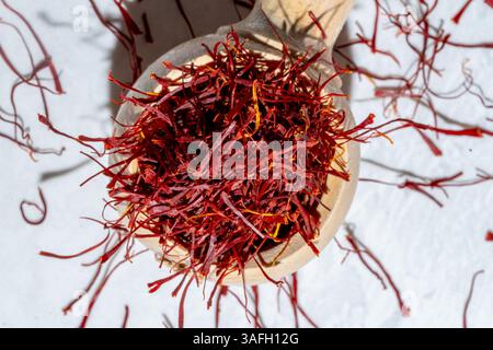Rote Safranfäden im Holzlöffel, die auf weißem Marmorhintergrund verschüttet werden, hell und luftig mit Sonnenlicht und Schatten, Ansicht von oben nach unten Stockfoto