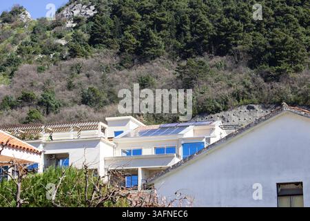 Weißes Öko-Haus mit Solarpaneelen und blauen Fensterrahmen in der Nähe der Küstenklippe. Nachhaltige Architektur in Montenegro Stockfoto