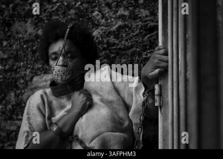 Schwarze Frau verkleidet als Sklave, mit einer eisernen Maske an Mund und Hals angekettet, an einem eisernen Zaun festgehalten. Sklaverei in Brasilien. Darstellung des Stockfoto