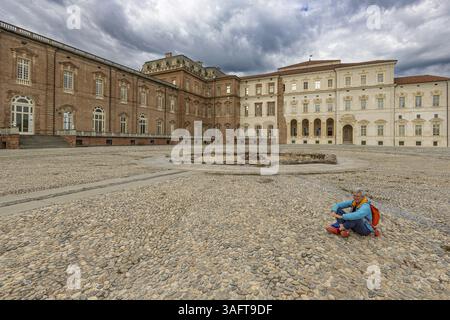 Frau, die auf einem Kieselplatz vor einem großen Gebäude sitzt, Palast Venaria reale, Residenzen des Hauses Savoyen, Turin, Piemont, Italien, Euro Stockfoto