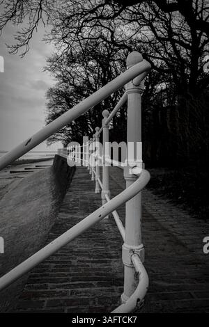 Ein Geländer entlang eines Kopfsteinpflasterpfads an der Küste, schwarz-weiß, Nordseestrand 26316 Varel Dangast-Deutschland Stockfoto