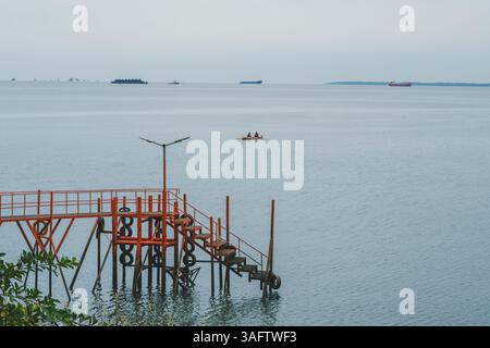 Eine friedliche Küstenlandschaft mit einem markanten roten Pier, der ins Meer führt, und einem kleinen Boot mit zwei Figuren in der Ferne. Stockfoto