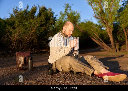Hispanischer Reisender, der bei Sonnenaufgang am Ufer eines Baches sitzt und die ersten Sonnenstrahlen empfängt, während er Mate trinkt, ein typisches argentinisches Getränk. El Stockfoto