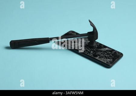 Smartphone mit zerbrochenem Glasbildschirm und Hammer auf blauem Hintergrund. Stockfoto
