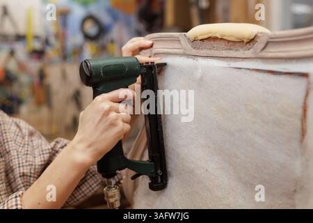 Nahaufnahme der Hände mit einem pneumatischen Hefter zum Befestigen der Polsterung an einem Vintage-Sessel während der Polsterarbeiten. Stockfoto
