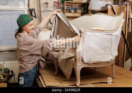 Lächelnde weibliche Polsterin dehnt Stoff über Vintage-Sofahmen aus, während sie Möbel in der Werkstatt restauriert. Stockfoto