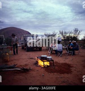 Outback-Campszene in Australien, vermutlich in der Nähe des Uluru (Ayers Rock), der im Hintergrund erkennbar ist. Eine Gruppe von Reisenden hat ihr Camp Handschuh in der Wüste aufgeschlagen und sitzen entspannt an einem Klapptisch. Im Vordergrund brennt ein Lagerfeuer, auf dem mehrere Kochtöpfe erhitzt werden. Ein Geländewagen, ausgestattet für Expeditionen, stehen bereit, während ein Mann mit Cowboyhut und Outdoor-Kleidung durch das Lager geht. Die Szene vermittelt das Gefühl von Abenteuer, Freiheit und der rauen Schönheit des australischen Outbacks, um 1985. Stockfoto