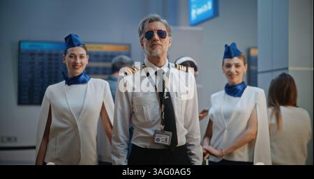 Porträt eines professionellen Flugzeugpiloten und zwei wunderschöner Flugbegleiter in eleganten Uniformen, die lächelnd in die Kamera schauen. Flugbesatzungsmitglieder im überfüllten internationalen Flughafenterminal. Mittlere Aufnahme. Stockfoto
