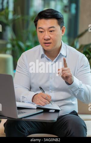 Junger chinesischer Geschäftsmann, der sich in der Hotellobby auf einem Laptop aufschreibt oder Notizen macht, während er an einem Online-Videokurs oder einem Büromeeting teilnimmt. Selbstbewusster Manager im Bereich T-Shirt-Support, der Webcam ansieht Stockfoto