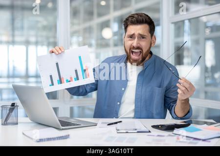 Ein frustrierter Geschäftsmann an seinem Schreibtisch zeigt schlechte finanzielle Ergebnisse auf einem Diagramm. Der Mann hält seine Brille und sieht verzweifelt aus. Stockfoto