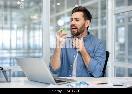 Ein Mann in einem Büro, der eine Halsschmerzen hat, benutzt ein Kehlspray, um bei der Arbeit an seinem Laptop zu lindern. Stockfoto