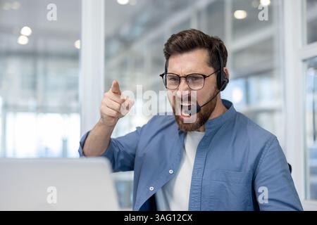 Ein wütender Kundendienstmitarbeiter schreit, während er frustriert mit dem Finger auf den Computerbildschirm vor sich zeigt Stockfoto