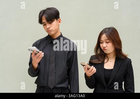 Porträt eines jungen asiatischen Paares, das bei der Benutzung des Mobiltelefons eine Geschäftsuniform vor einem schlichten Hintergrund trägt Stockfoto