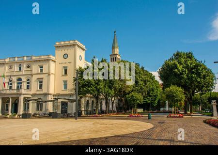 Piazza della Repubblica in Monfalcone, Friaul, Italien. Platz mit Springbrunnen, Blumenbeeten von Impatiens. Links: Neoklassizistisches Rathaus, Glockenturm der Kathedrale Stockfoto