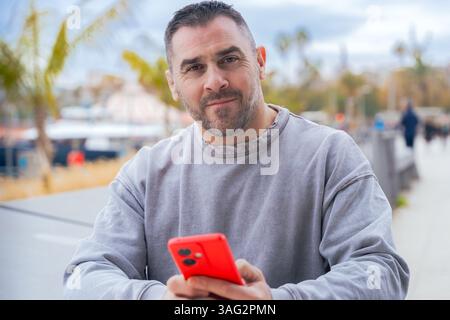 Mann tippt auf rotem Smartphone, während er draußen in der Nähe von Palmen und Hafen sitzt Stockfoto