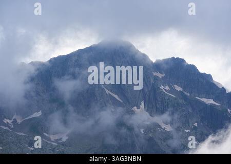 Wolken, die über steile, felsige Berghänge mit Schneeflächen und dunklen Granitwänden schweben Stockfoto