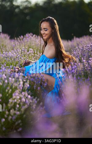 Junge Frau in blauem Kleid posiert selbstbewusst in einem blühenden Lavendelfeld während Sonnenuntergang Stockfoto