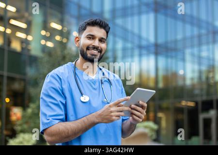 Ein indischer Arzt lächelt, während er ein Tablet hält und möglicherweise die Patientendaten im Freien vor dem Hintergrund des Gebäudes überprüft. Stockfoto