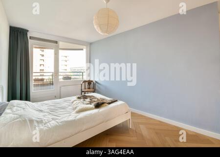 Ein helles und luftiges, modernes Schlafzimmer mit weißem Bett, weichen Texturen und Tageslicht. Die beruhigende blaue Wand und das schlichte Dekor schaffen eine ruhige Atmosphäre. Stockfoto