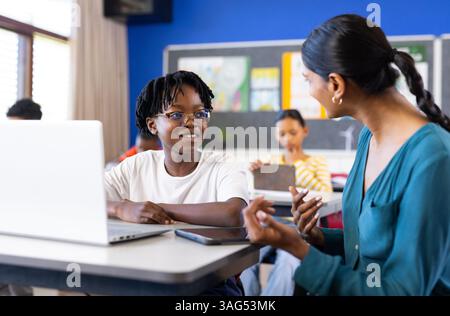 In der Schule hilft eine indische Lehrerin der Schülerin mit Laptop im Klassenzimmer, lächelt und redet Stockfoto