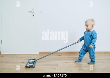 In einer gemütlichen Innenumgebung wischt ein fröhliches Kind in blauen Overalls gerne den Boden und lernt wertvolle Lektionen über Verantwortung und Lebensfähigkeiten Stockfoto