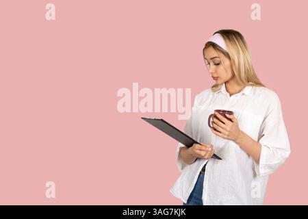 Seitenporträt einer schönen blonden Geschäftsfrau mit Klemmbrett. Kaffeetasse halten, denken, lesen. In formellem Outfit über Rosa stehen Stockfoto