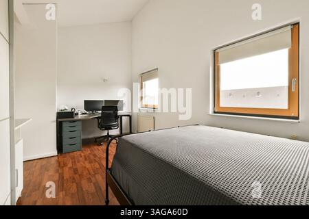 Ein geräumiges und gut beleuchtetes minimalistisches Schlafzimmer mit Bett, Schreibtisch und Stuhl. Das Design betont Einfachheit und Funktionalität mit natürlichem Licht, das durch das Fenster strömt. Stockfoto