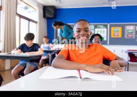 Lächelnder Junge im Klassenzimmer mit offenem Notizbuch, Schüler und indische Lehrerin im Hintergrund Stockfoto