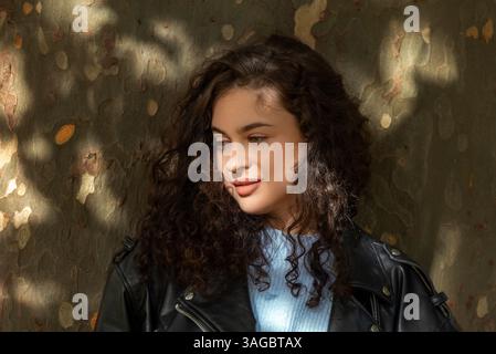 Eine junge Frau mit dunklen lockigen Haaren, die sich auf einen Baumstamm stützt. Porträt einer Millennial-Frau Stockfoto