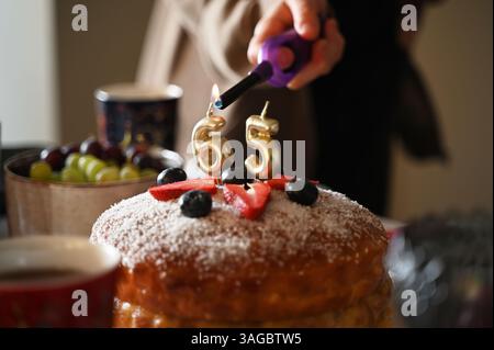 Ein festliches Nahaufnahme-Bild, das den Moment festmacht, in dem Kerzen auf einer Geburtstagskuche zum 65. Geburtstag angezündet werden. Der Kuchen ist mit frischen s dekoriert Stockfoto