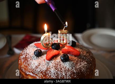 Ein festliches Nahaufnahme-Bild, das den Moment festmacht, in dem Kerzen auf einer Geburtstagskuche zum 65. Geburtstag angezündet werden. Der Kuchen ist mit frischen s dekoriert Stockfoto