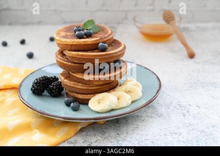Süße und flauschige, gestapelte Pfannkuchen mit Bananen und Beeren. Mit Honig auf blauen Tellern gegen hellgraue Tischplatte mit gelbem Tuch Stockfoto