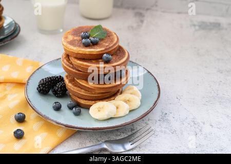 Süße und flauschige, gestapelte Pfannkuchen mit Bananen und Beeren. Mit Honig auf blauen Tellern gegen hellgraue Tischplatte mit gelbem Tuch Stockfoto