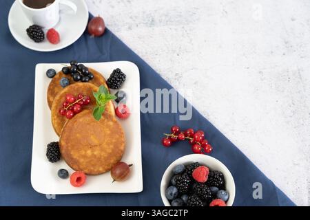 Frisch gestapelte Pfannkuchen mit verschiedenen Beeren auf weißer Platte auf einem blauen diagonalen Tuch auf grauem Hintergrund Stockfoto