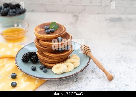 Süße und flauschige, gestapelte Pfannkuchen mit Bananen und Beeren. Mit Honig auf blauen Tellern gegen hellgraue Tischplatte mit gelbem Tuch Stockfoto