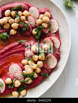 Lebendiger Rote-Bete-Hummus auf einer rustikalen Keramikplatte serviert, garniert mit Kichererbsen, frischen Rettichscheiben, gehackten Kräutern und einem Hauch Olivenöl. Stockfoto