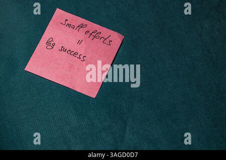 Ein rosafarbener Haftnotizblock mit den Worten „kleine Anstrengungen, großer Erfolg“ wird auf einer strukturierten grünen Oberfläche platziert. Dieser Vermerk vermittelt eine positive Botschaft über achi Stockfoto