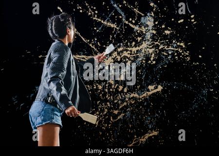 Künstler in Jeansshorts, die abstrakte Splatter-Kunst auf dunkler Wand kreieren. Künstlerisch, kreativ, Malerei, Ausdruck, zeitgenössisch, Kreativität Stockfoto