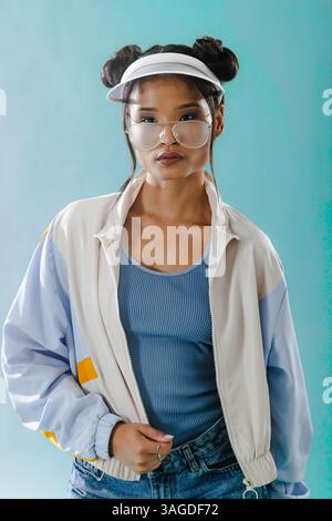 Junge Frau in Retro-Jacke und Visier posiert selbstbewusst vor blauem Hintergrund. Mode, Style, Vintage, trendig, lebendig Stockfoto