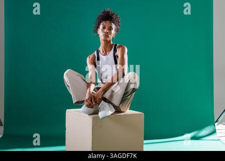 Das Model sitzt im Studio auf Cube, trägt ein lässiges Outfit und sieht selbstbewusst aus. Pose, Kleidung, trendig, Porträt, Stil Stockfoto