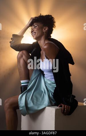 Afroamerikanerin lacht fröhlich, sitzt auf einem Hocker in stilvollem Grunge-Outfit. Mode, Urban, lässig, Trend, Porträt, Lifestyle Stockfoto