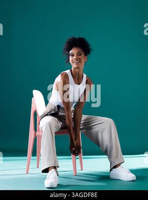Modebewusste Frau im lässigen Outfit, die im Studio selbstbewusst auf dem Stuhl sitzt. Stil, Selbstvertrauen, trendig, modellierend, schick Stockfoto