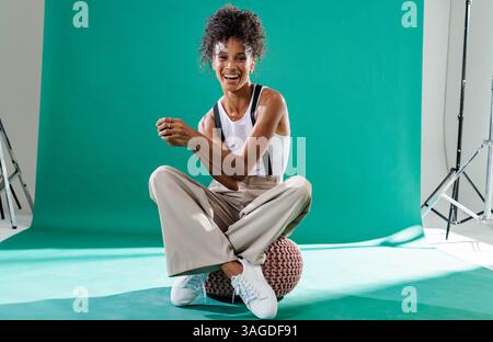 Lächelnde Frau, die im Studio auf dem Hocker sitzt, lässiges Outfit und Turnschuhe trägt. Mode, Stil, Bekleidung, Schuhe, Porträt, komfortabel Stockfoto