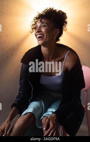 Afroamerikanerin lacht fröhlich in stilvollem Outfit, sitzt auf einem Stuhl. Mode, Lifestyle, Glück, Eleganz, Sitzen, Ausdruck Stockfoto
